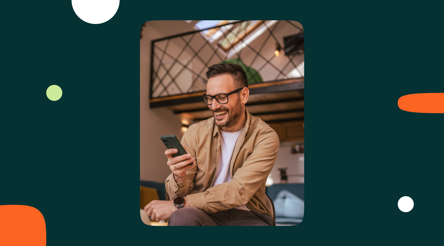 Man with glasses smiling at his smartphone while sitting in a modern loft-style interior, representing a customer engaging with a conversational AI interface on mobile.